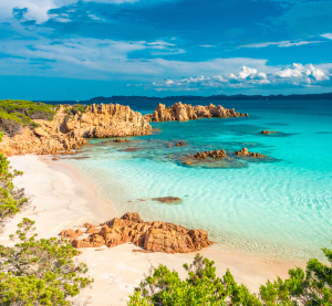 Traumhafter Sandstrand mit t&uuml;rkisblauem Wasser und r&ouml;tlichen Felsen auf Sardinien, umgeben von gr&uuml;ner Mittelmeer-Vegetation unter blauem Himmel mit Wolken.