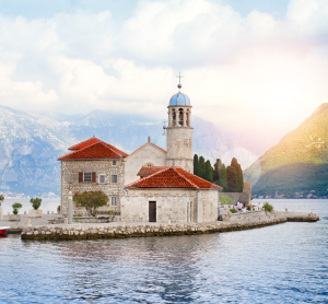 Kleine historische Kirche mit rotem Ziegeldach auf einer Insel in der Bucht von Kotor in Montenegro, umgeben von ruhigem Wasser und Bergen im weichen Sonnenlicht.