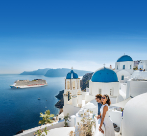 Paar blickt von den wei&szlig;en H&auml;usern mit blauen Kuppeln in Santorini auf ein Kreuzfahrtschiff im tiefblauen Meer.