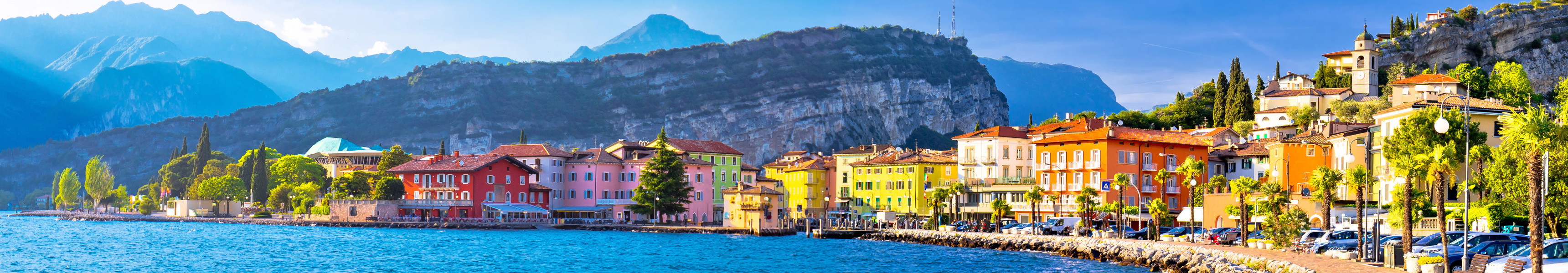 Bunte H&auml;user am Ufer des Gardasees mit Bergkulisse im Hintergrund bei sonnigem Wetter.