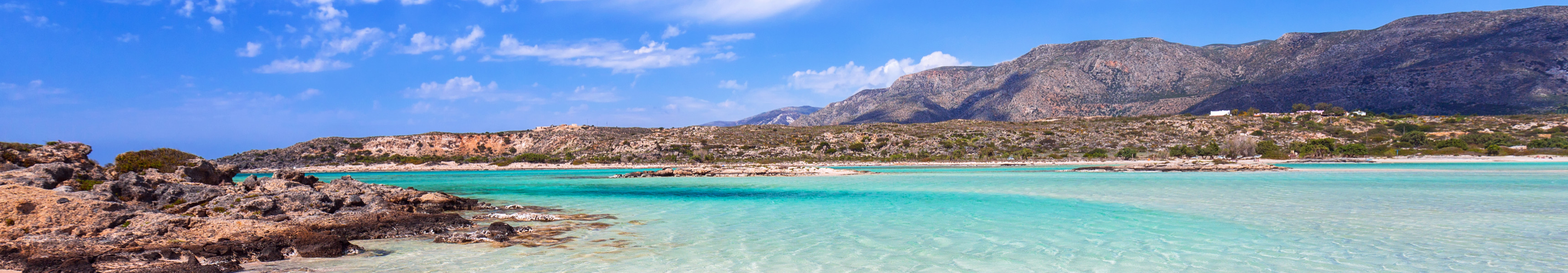 Traumhafte Lagune von Elafonissi auf Kreta mit rosa schimmerndem Sand, t&uuml;rkisblauem Wasser und felsiger K&uuml;ste vor einer Bergkulisse unter blauem Himmel mit wei&szlig;en Wolken.