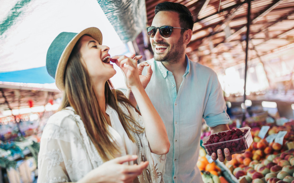 Paar steht an einem Marktstand und die Frau probiert eine Himbeere, die ihr der Mann reicht.