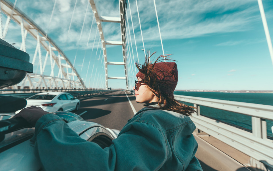 Person mit M&uuml;tze und Sonnenbrille lehnt sich aus einem fahrenden Auto auf einer Br&uuml;cke, im Hintergrund flie&szlig;t der Verkehr und das Meer ist sichtbar.