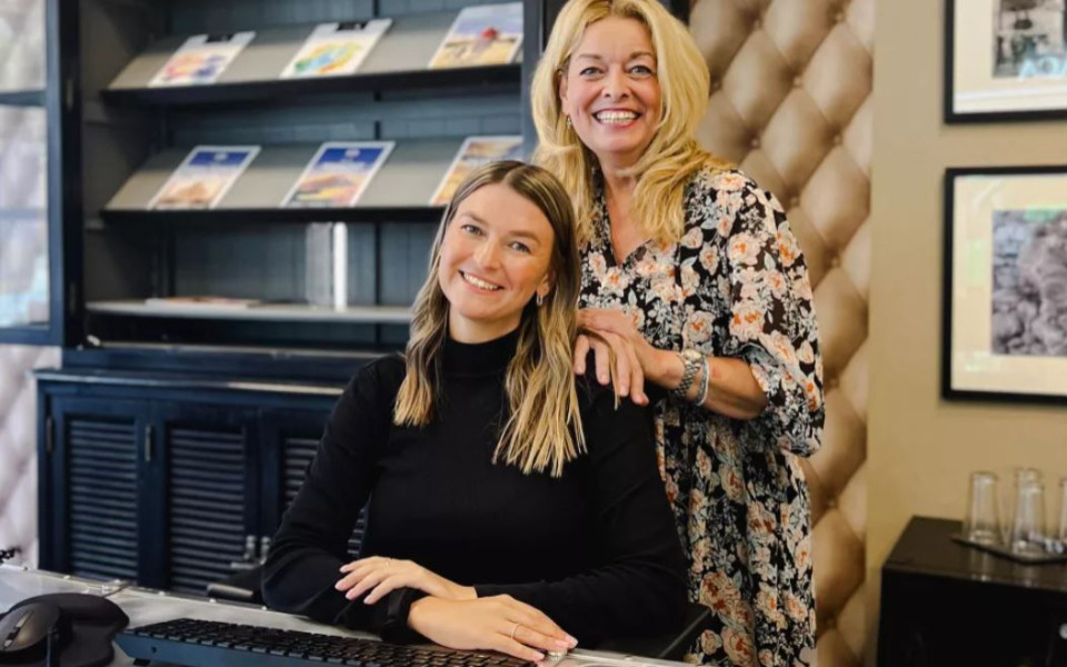 Zwei Reiseb&uuml;romitarbeiterinnen im B&uuml;ro, eine sitzt am Schreibtisch mit Tastatur, die andere steht dahinter vor einem Regal mit Reisekatalogen.