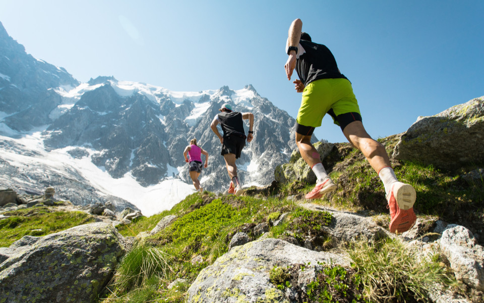 Drei Trailrunner, zwei M&auml;nner und eine Frau, laufen an einem hei&szlig;en, hellen Sommertag einen steilen Pfad in den Bergen der Alpen hinauf.  