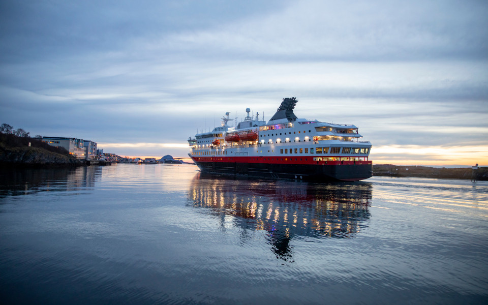 Ein Schiff der Hurtigruten f&auml;hrt bei Abendd&auml;mmerung durch einen ruhigen Fjord in Norwegen. 