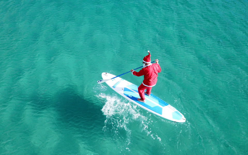 Als Weihnachtsmann verkleideter Mann paddelt auf einem Surfbrett im t&uuml;rkisblauen Meer in Australien.