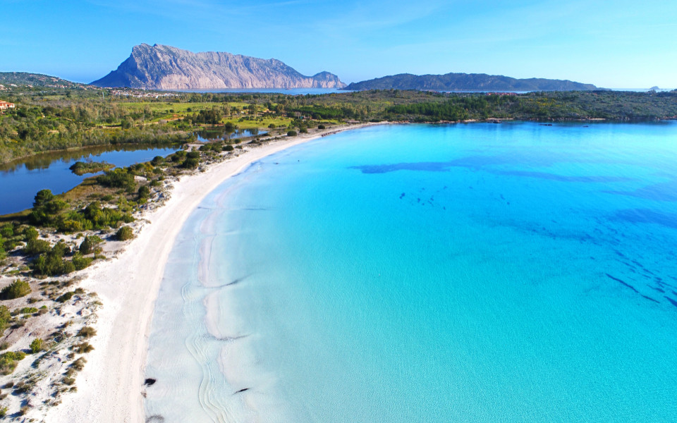 Blick auf einen Strand mit kristallblauem Meer und weißem Sandstrand, dahinter ist ein Wald und Gebirge.